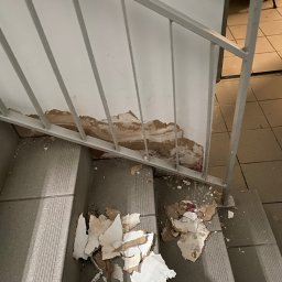 Dominik Mroczkowski - Uszkodzona ściana na klatce schodowej z odpadającym tynkiem i farbą, widoczne schody i metalowa barierka.