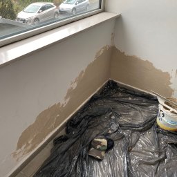 Dominik Mroczkowski - Przygotowanie ściany do malowania: częściowo nałożona warstwa szpachli, wiadro z gotową masą szpachlową, szpachelki na folii ochronnej, widok z okna na zaparkowane samochody.