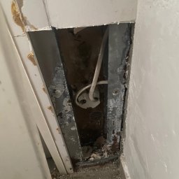 Dominik Mroczkowski - Otwór w ścianie odsłaniający instalację elektryczną z widocznymi przewodami i puszką podtynkową, zniszczone wykończenie ściany i widoczne ślady korozji.