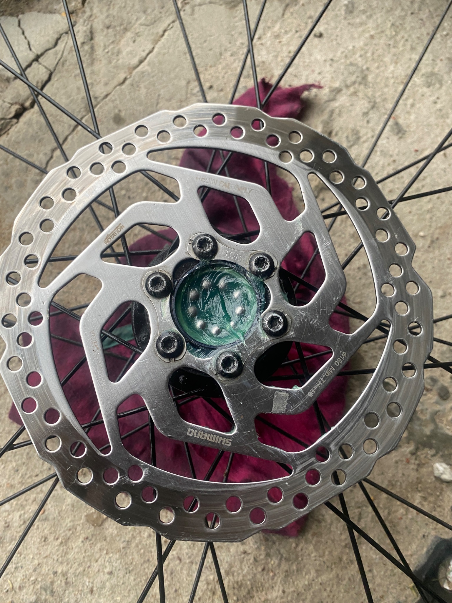 Tarcza hamulcowa roweru marki Shimano, z widocznymi śladami smaru i zabrudzeniami, na tle szprych i fragmentu fioletowej szmatki.