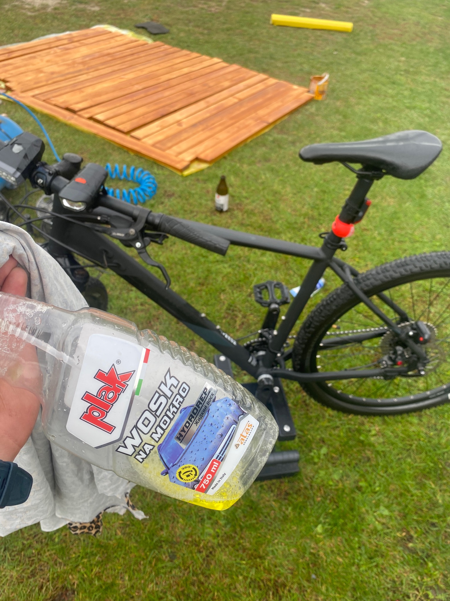 Czarny rower górski na trawie obok świeżo pomalowanych desek; w pierwszym planie butelka wosku samochodowego 'Plak Wosk na Mokro' trzymana w dłoni.