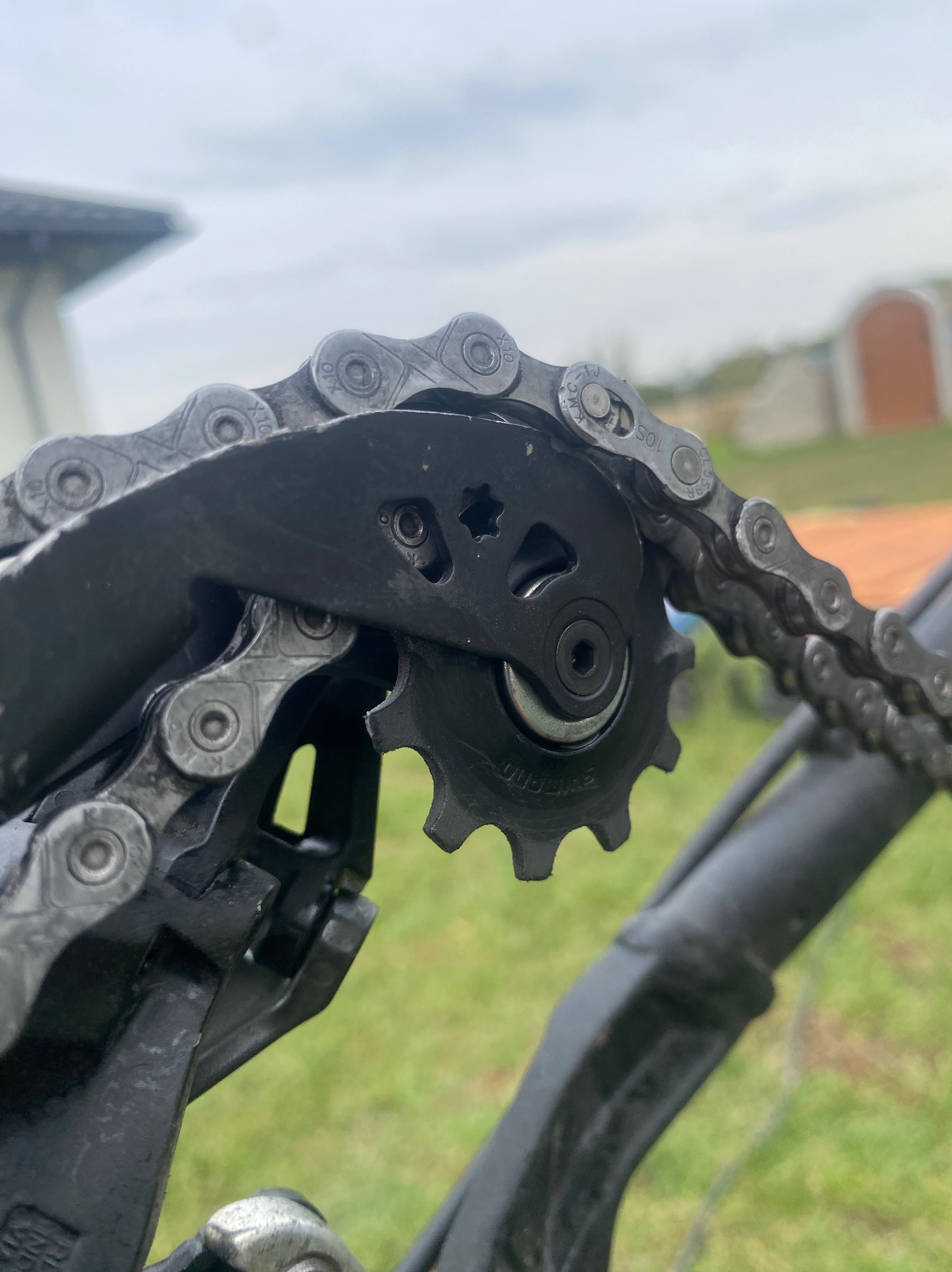 Detail czarnej przerzutki rowerowej z łańcuchem na tle zielonej trawy i zarysu budynku w pochmurny dzień.