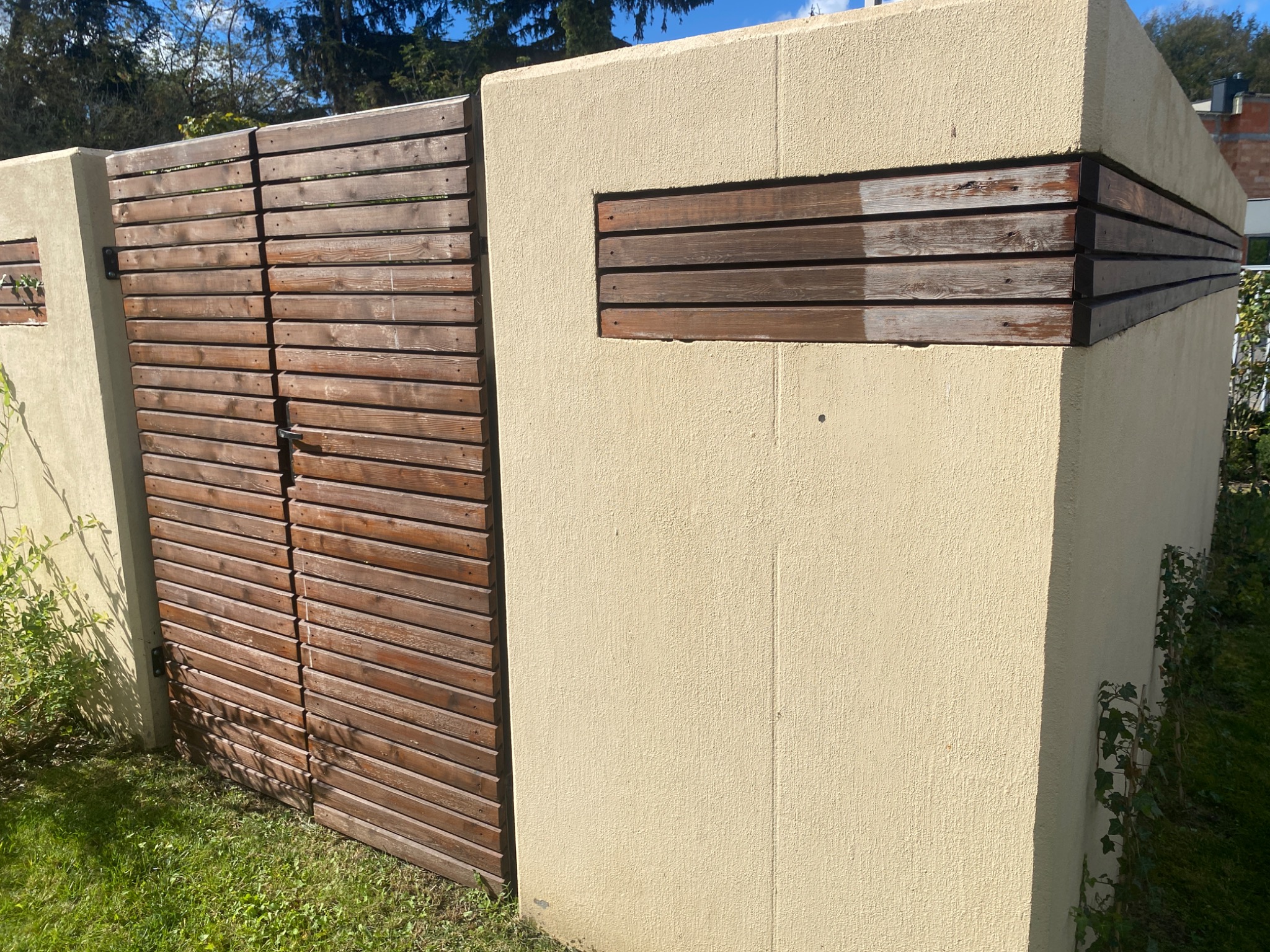 Nowoczesna brama wjazdowa z poziomych desek, wkomponowana w beżowy, otynkowany mur w Świerczach. Fragment muru posiada dekoracyjne wstawki z drewna.