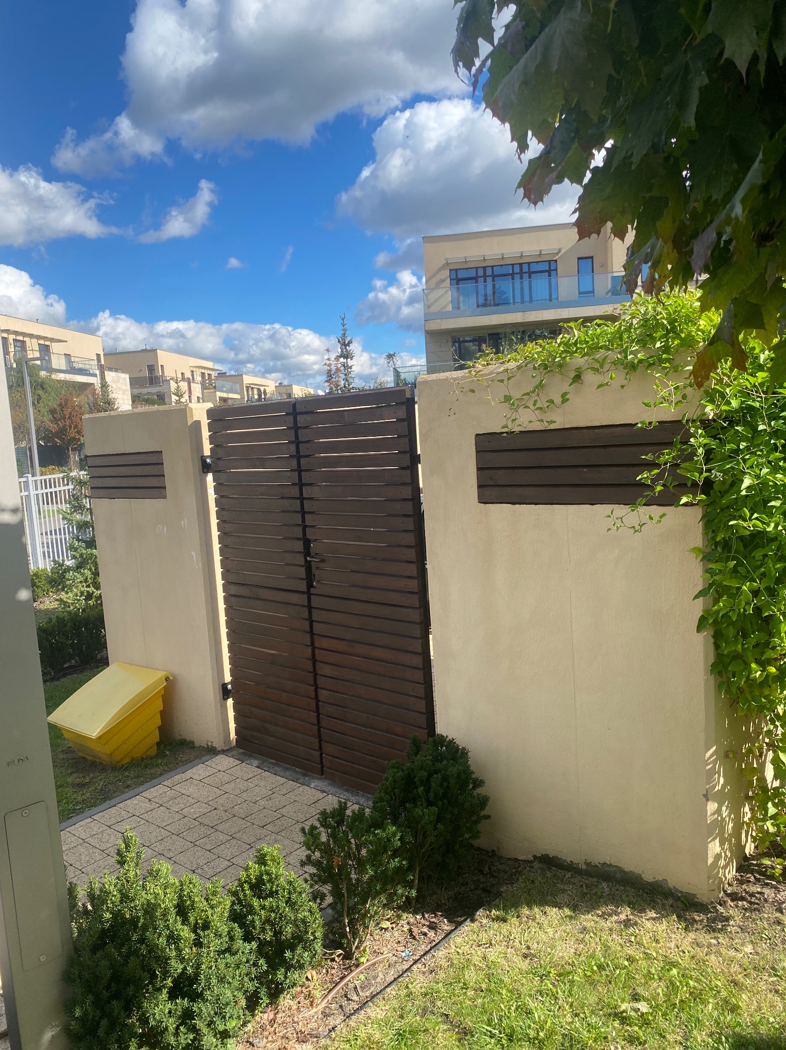 Nowoczesna brama z poziomych desek w jasnym ogrodzeniu, widok z zewnątrz na tle budynków mieszkalnych i błękitnego nieba z chmurami, z elementami zieleni.