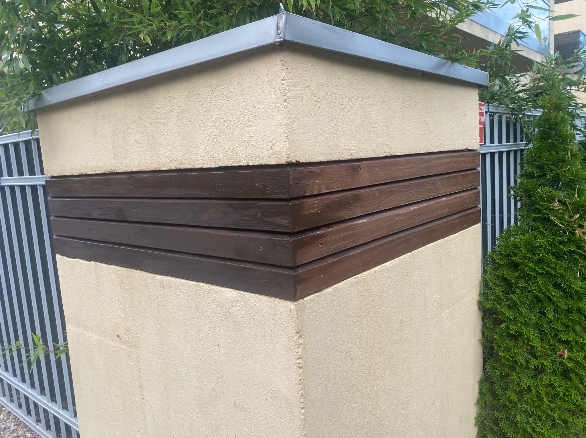 Wykończony fragment ogrodzenia z beżowego tynku i poziomych, ciemnobrązowych desek, zwieńczony metalowym daszkiem, na tle zieleni i metalowego ogrodzenia.