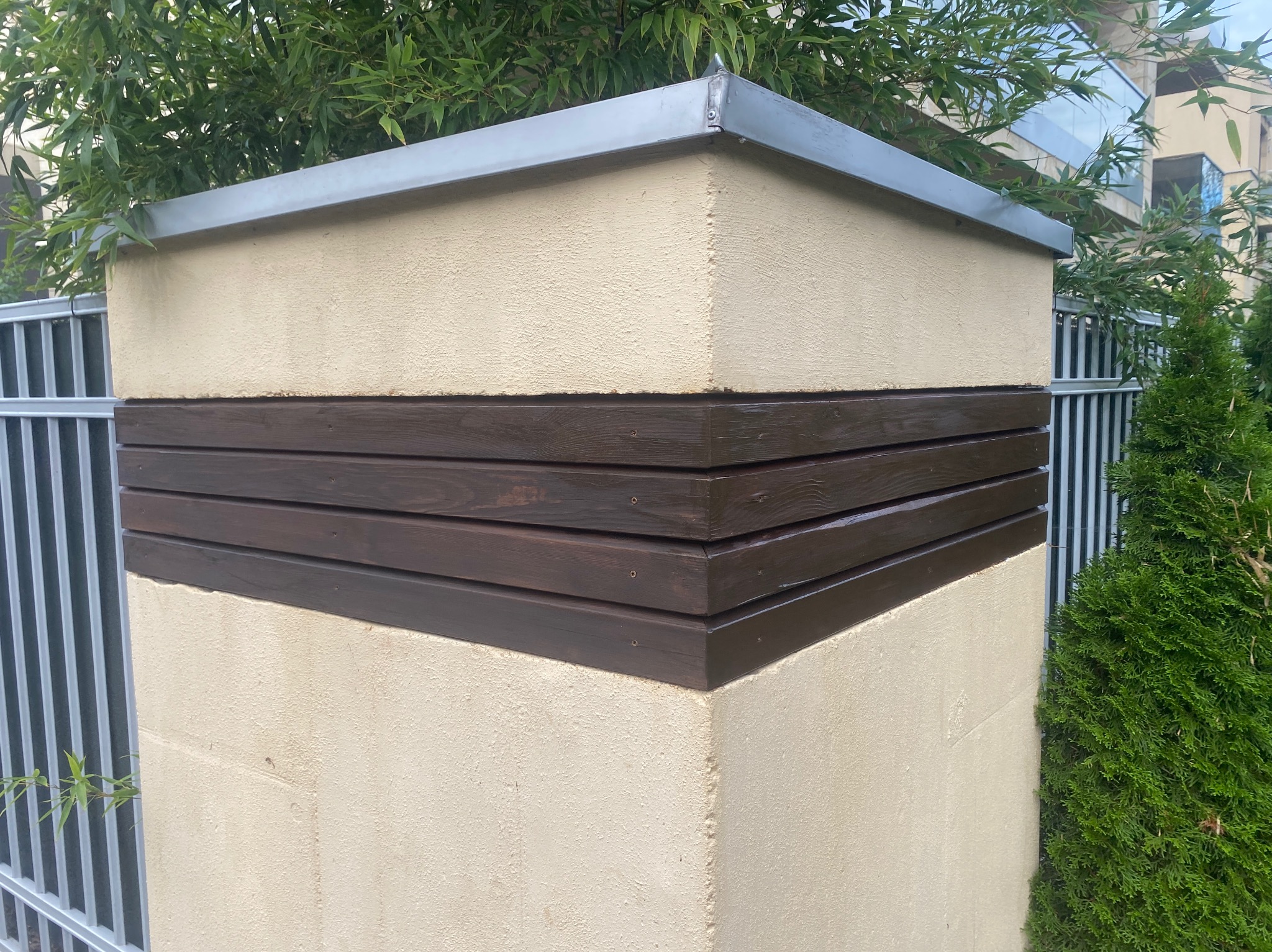 Fragment ogrodzenia z beżowego tynku z ciemnobrązowymi, drewnianymi panelami i metalowym daszkiem, otoczony zielenią.