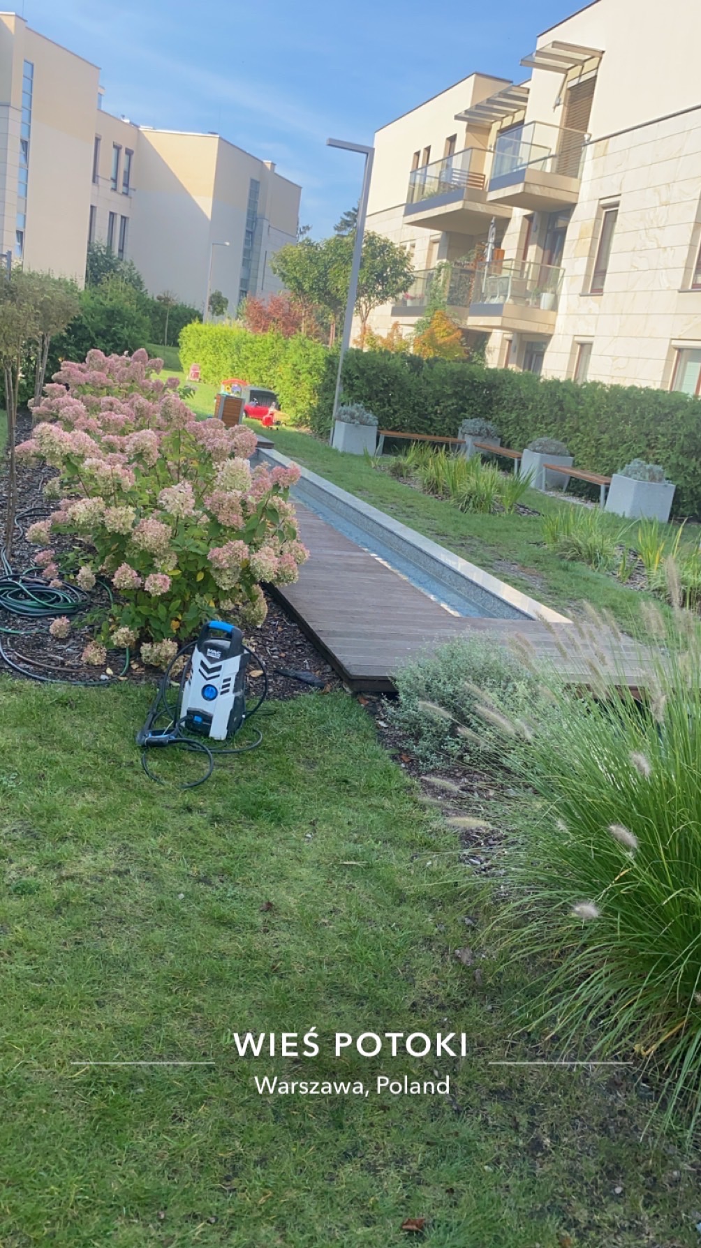 Myjka ciśnieniowa stojąca na trawie obok krzewu hortensji, drewnianej kładki i niskiego murku w otoczeniu nowoczesnej zabudowy osiedlowej.
