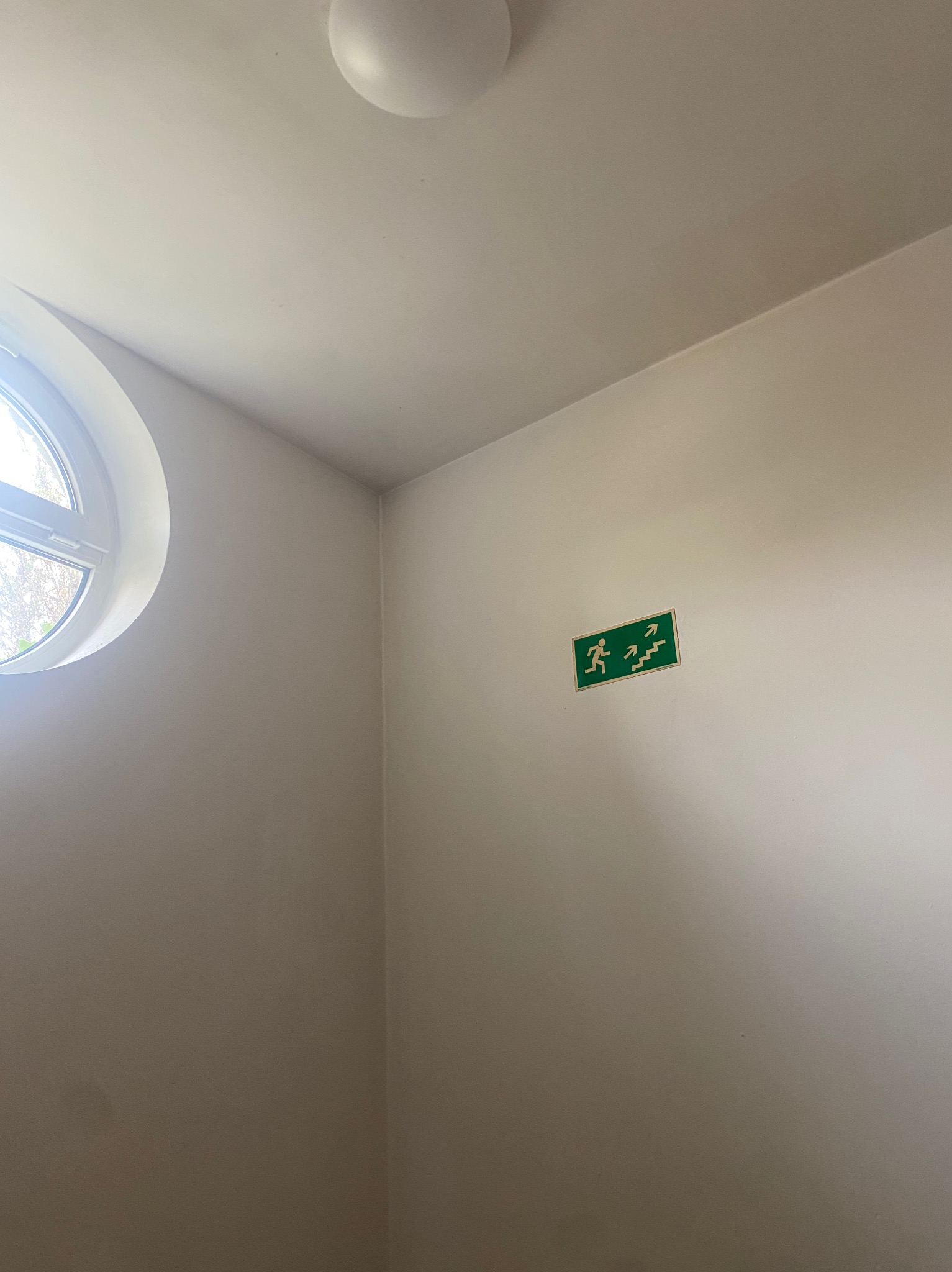 Wnętrze klatki schodowej z okrągłym oknem i zielonym znakiem ewakuacyjnym na ścianie, świeżo po malowaniu na jasny odcień.