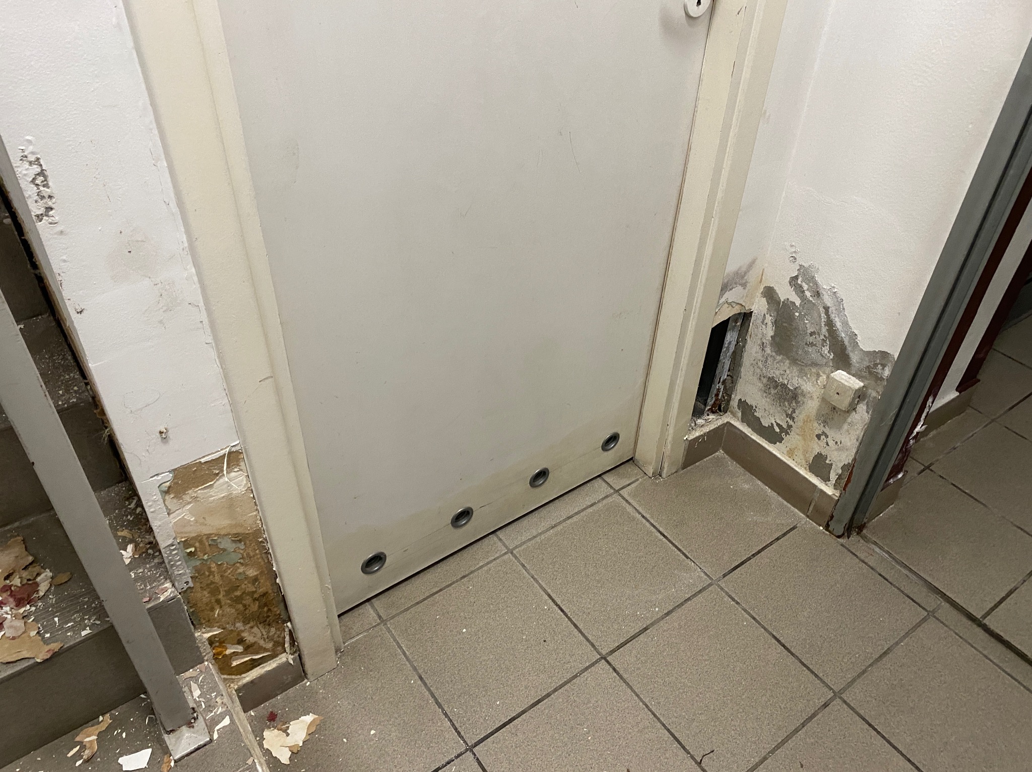 Uszkodzona dolna część futryny i ściany przy drzwiach wejściowych, widoczne ślady wilgoci i odpadający tynk, odsłonięta warstwa izolacyjna, brudna podłoga z płytek.