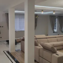 Przestronny salon z narożną sofą w odcieniach beżu, marmurowe schody z podświetleniem LED, parkiet w kolorze naturalnego drewna, dekoracyjne żyrandole i minimalistyczna choinka, tworzące nowoczesne...