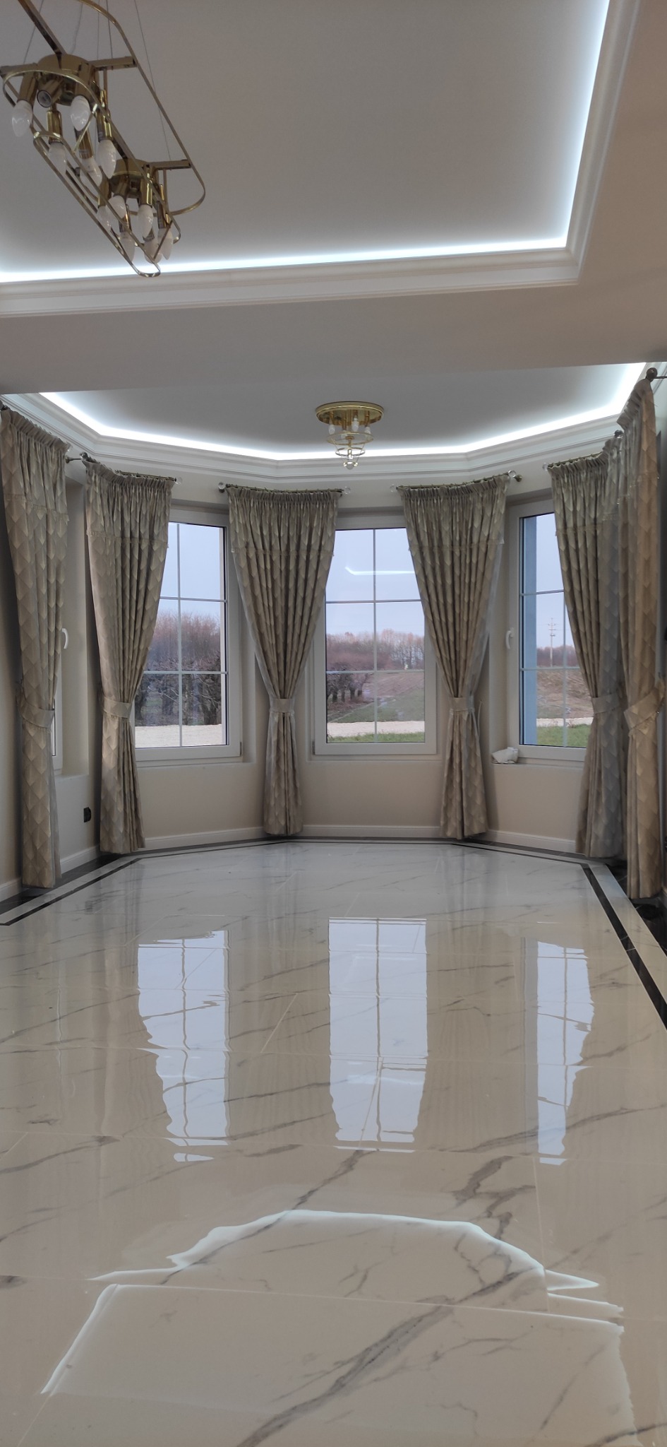 Przestronne wnętrze z błyszczącą, marmurową podłogą odbijającą światło z okien z beżowymi zasłonami; widoczne detale wykończeniowe sufitu oraz nowoczesne oświetlenie.