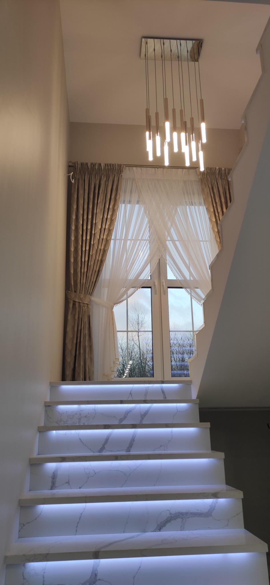 Schody z podświetleniem LED i marmurowym wzorem, okno z firanami i zasłonami w tle, nowoczesny żyrandol.