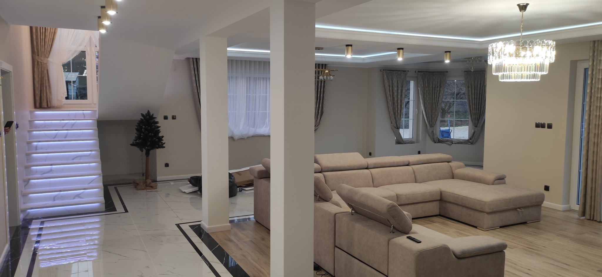 Przestronny salon z narożną sofą w odcieniach beżu, marmurowe schody z podświetleniem LED, parkiet w kolorze naturalnego drewna, dekoracyjne żyrandole i minimalistyczna choinka, tworzące nowoczesne...