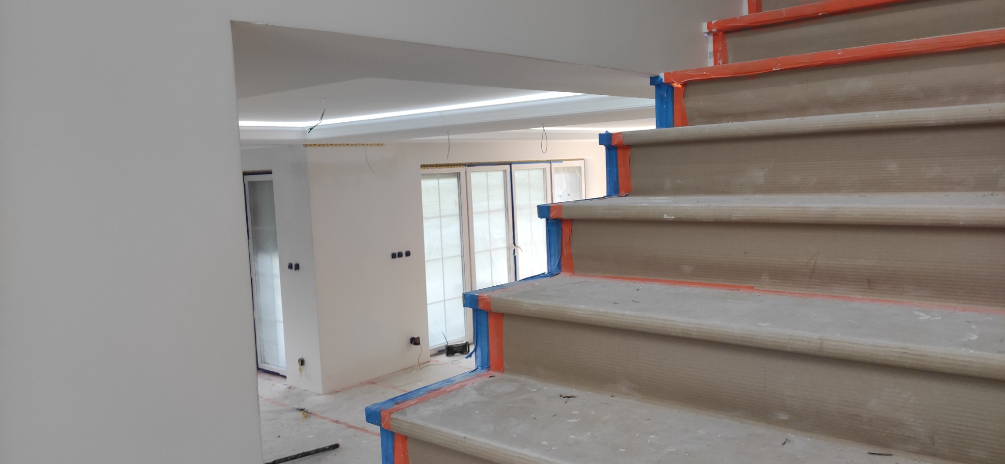 Schody zabezpieczone taśmą malarską podczas prac wykończeniowych w jasnym wnętrzu z oknami balkonowymi i oświetleniem LED.