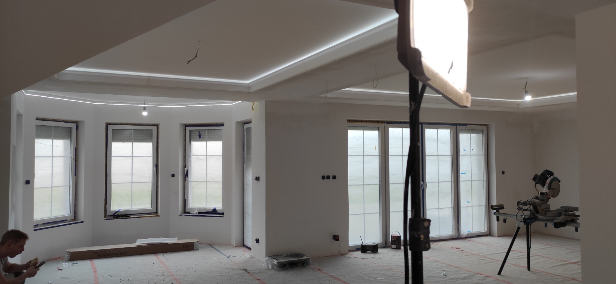 Wnętrze w trakcie prac wykończeniowych: widoczne okna zabezpieczone folią, listwy oświetleniowe na suficie, narzędzia pracy (pilarka) i pracownik w rogu pomieszczenia.