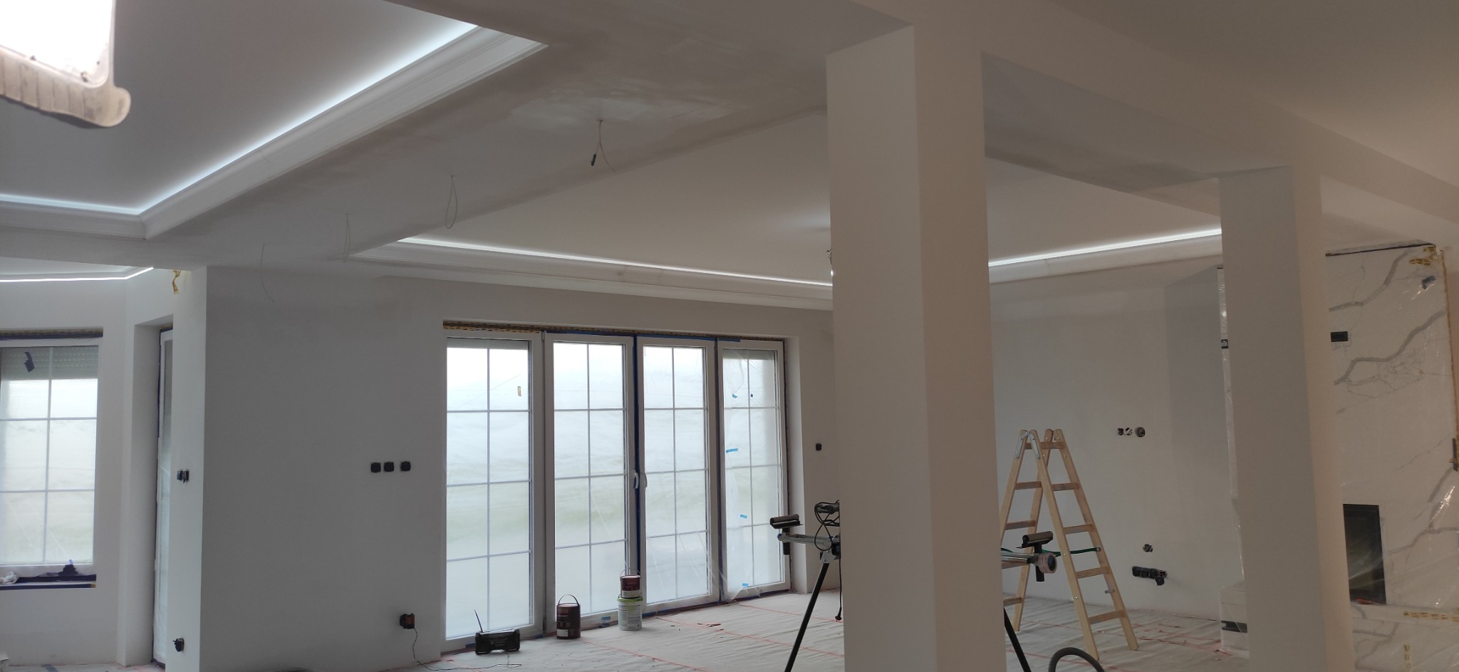 Wnętrze w trakcie wykończenia: ściany po szpachlowaniu i malowaniu na biało, widoczne listwy LED na suficie, okna zabezpieczone folią, drabina i narzędzia malarskie na podłodze.