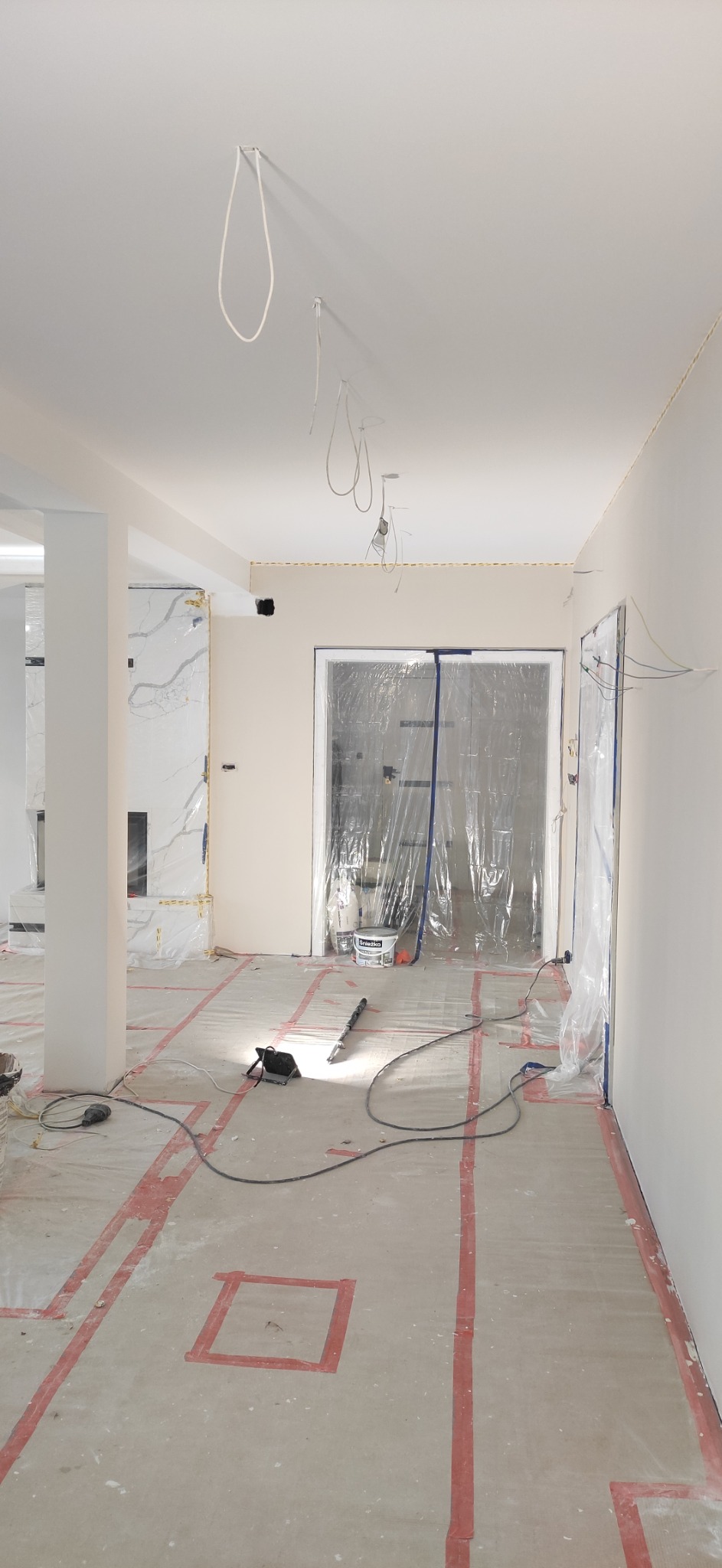 Przygotowania do prac wykończeniowych w jasnym wnętrzu z zabezpieczonymi folią drzwiami, widoczne przewody elektryczne zwisające z sufitu, oświetlenie robocze i taśmy zabezpieczające podłogę.
