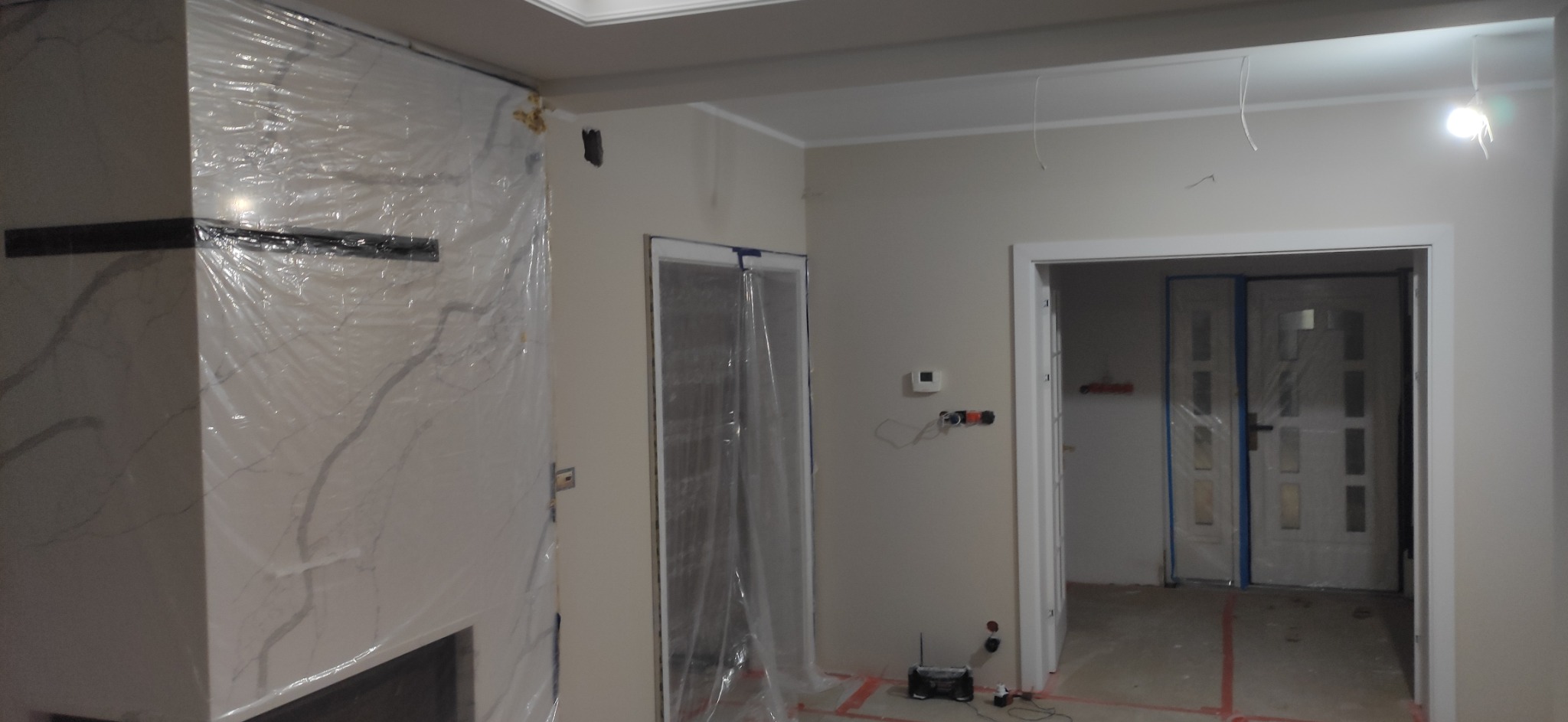 Pokój w trakcie remontu: ściany przygotowane do malowania, zabezpieczone folią drzwi i kominek z marmurowym wykończeniem, widoczne wystające kable i osprzęt elektryczny.