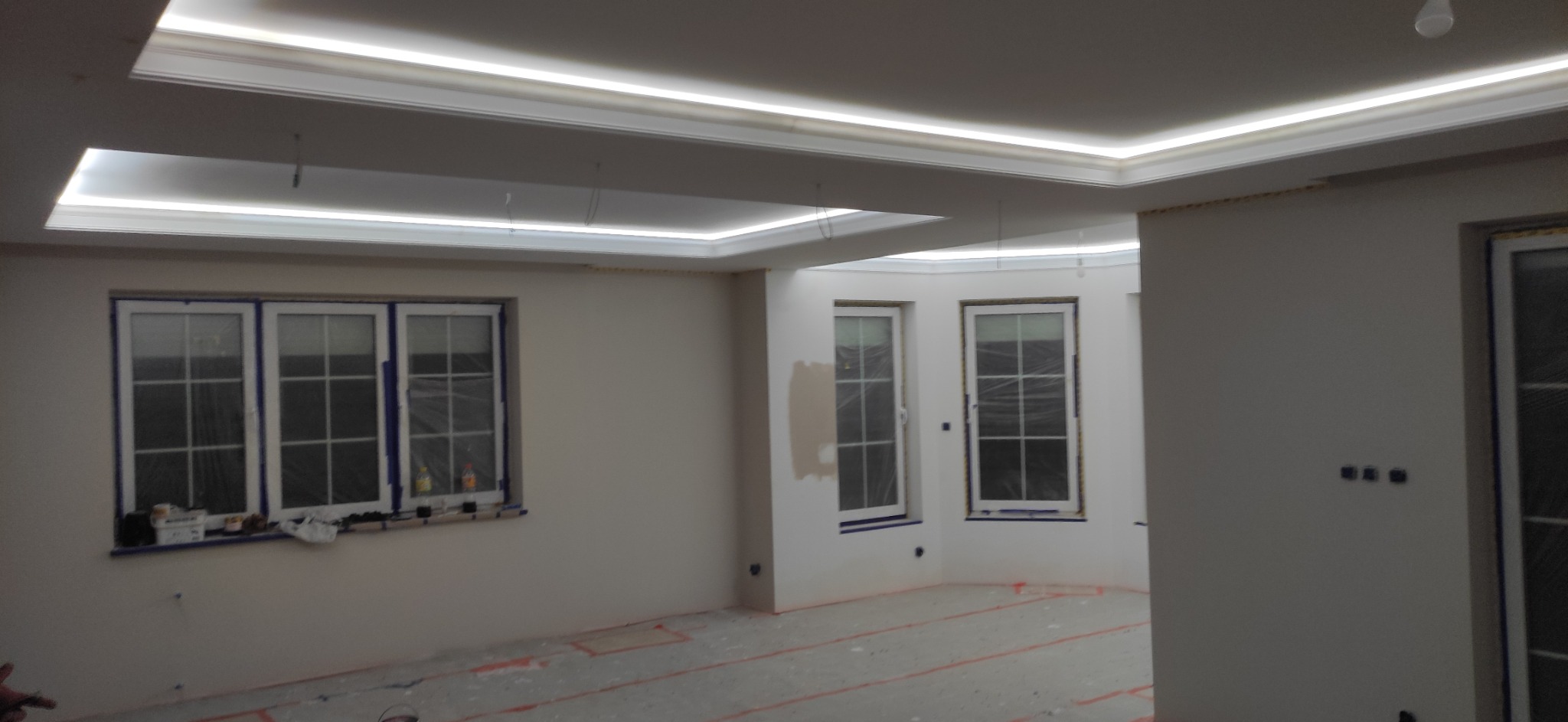Pokój w trakcie remontu z trzema oknami zabezpieczonymi folią, widoczne ślady po szpachlowaniu na ścianach i oświetlenie LED w suficie podwieszanym.