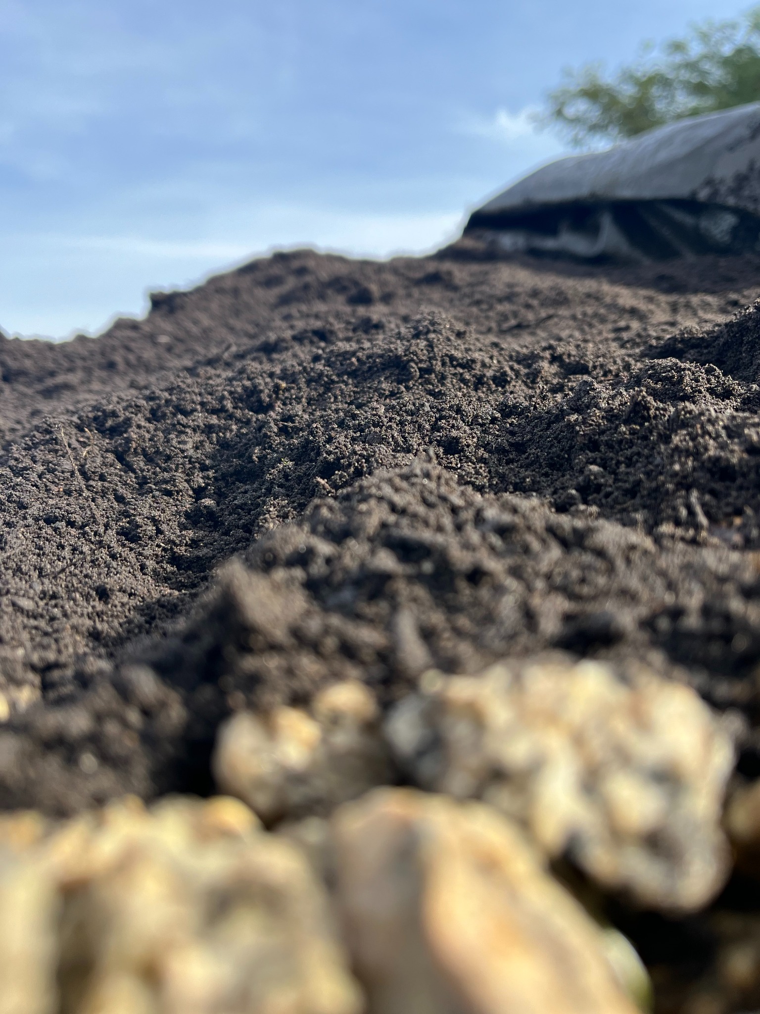 Kadr z bliska na pryzmę ciemnej ziemi ogrodowej z rozmytymi jasnymi kamieniami na pierwszym planie, pod błękitnym niebem.