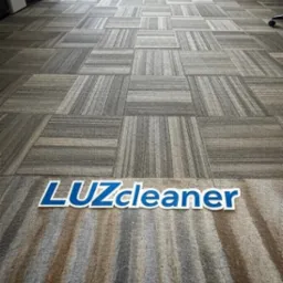 Wzorzysta wykładzina biurowa w odcieniach szarości z logo 'LUZcleaner' na pierwszym planie. Widok z perspektywy podłogi, w tle krzesła biurowe i okno.