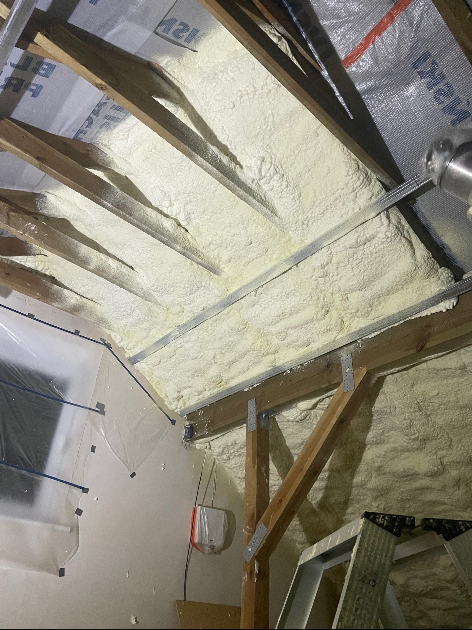 Poddasze z widocznymi krokwiami drewnianymi i metalowymi profilami, pokryte grubą warstwą jasnej pianki izolacyjnej; w rogu widoczna drabina i folia ochronna na ścianie.