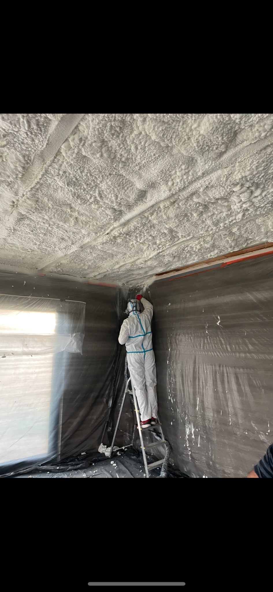 Pracownik w kombinezonie ochronnym na drabinie aplikuje piankę izolacyjną na sufit i ściany pomieszczenia zabezpieczonego folią.