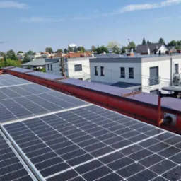 Panele słoneczne na czerwonym dachu z widoczną anteną satelitarną i kominem w tle, zabudowa jednorodzinna w tle.