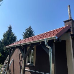 Dachowiec - Dom z nową, czerwoną dachówką i błyszczącymi, metalowymi rynnami, widok z dołu, drabina oparta o ścianę, błękitne niebo w tle.