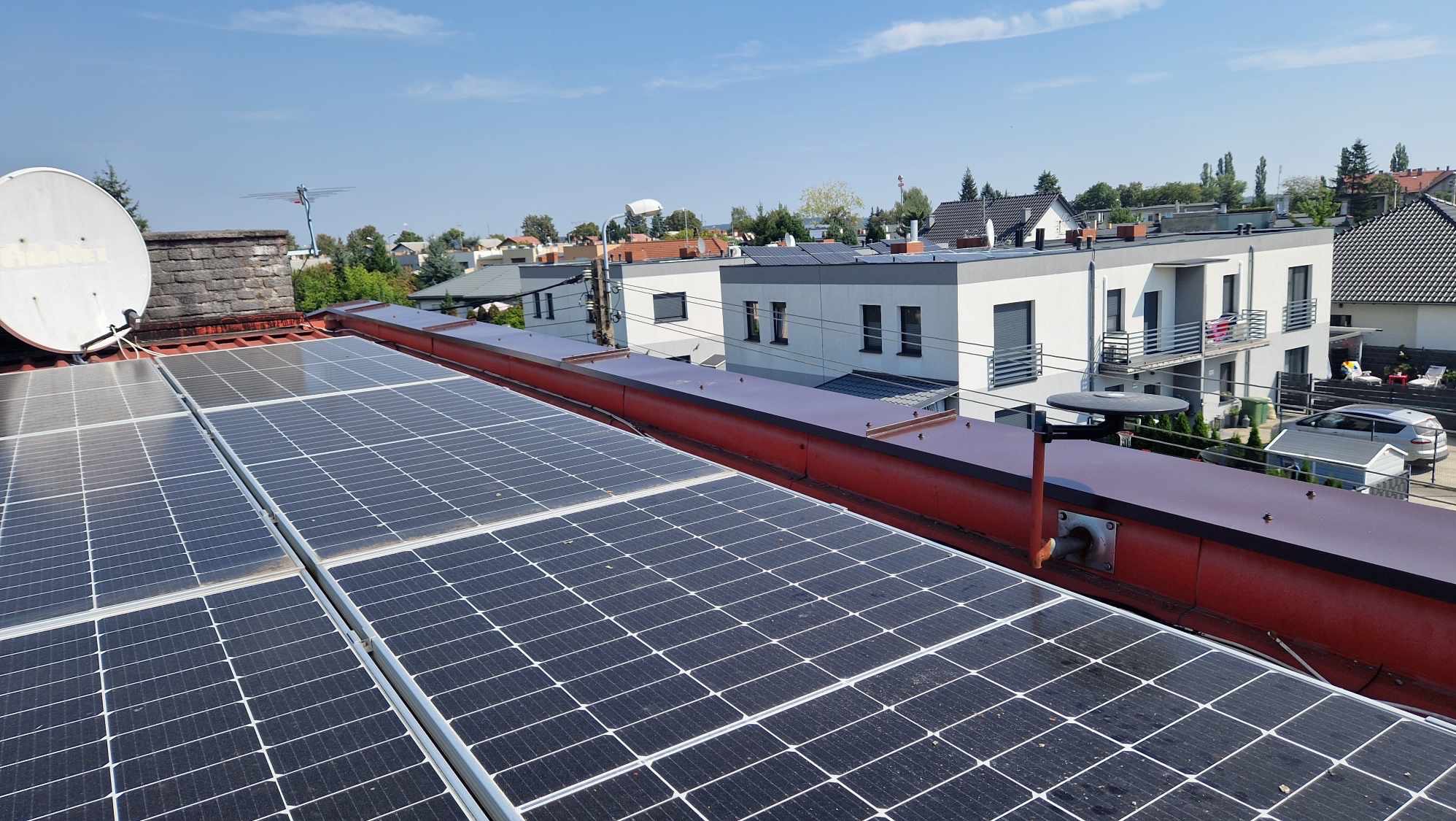 Panele słoneczne na czerwonym dachu z widoczną anteną satelitarną i kominem w tle, zabudowa jednorodzinna w tle.