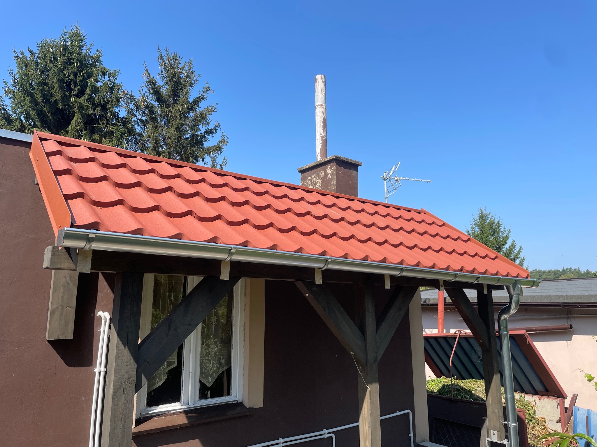 Dom z nowym, czerwonym dachem z blachodachówki, srebrne rynny, ciemnobrązowa elewacja, widoczny komin i antena telewizyjna, drzewa w tle, słoneczny dzień.