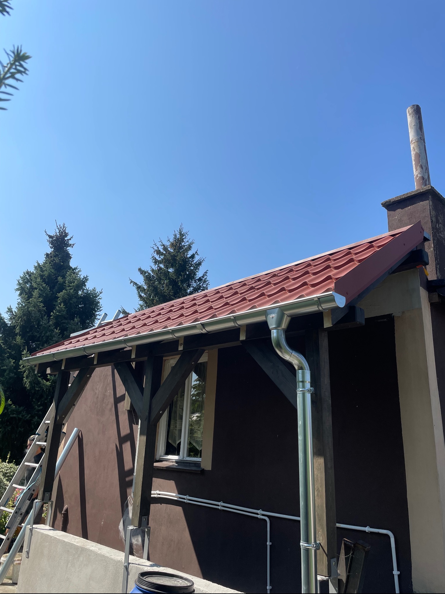 Dom z nową, czerwoną dachówką i błyszczącymi, metalowymi rynnami, widok z dołu, drabina oparta o ścianę, błękitne niebo w tle.
