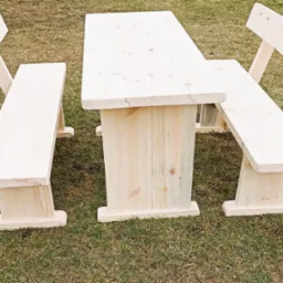 Surowy, drewniany stół piknikowy z dwiema ławkami, ustawiony na trawniku. Widoczne naturalne słoje drewna i solidna konstrukcja.