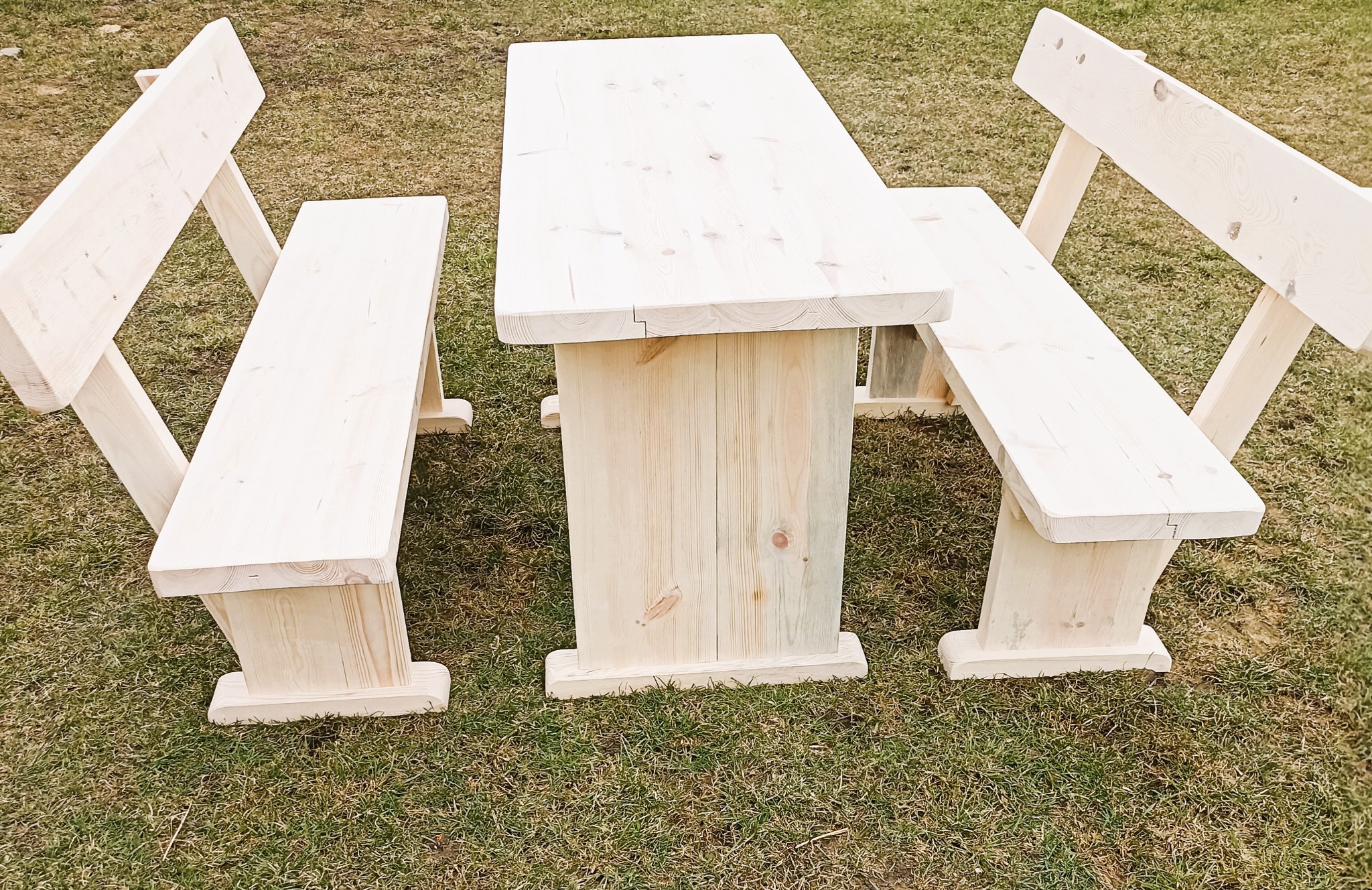 Surowy, drewniany stół piknikowy z dwiema ławkami, ustawiony na trawniku. Widoczne naturalne słoje drewna i solidna konstrukcja.