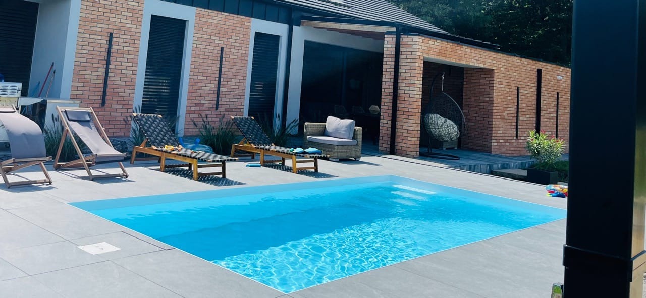 Prostokątny basen z błękitną wodą w ogrodzie przy nowoczesnym domu z cegły, obok leżaki i rattanowy fotel wiszący.