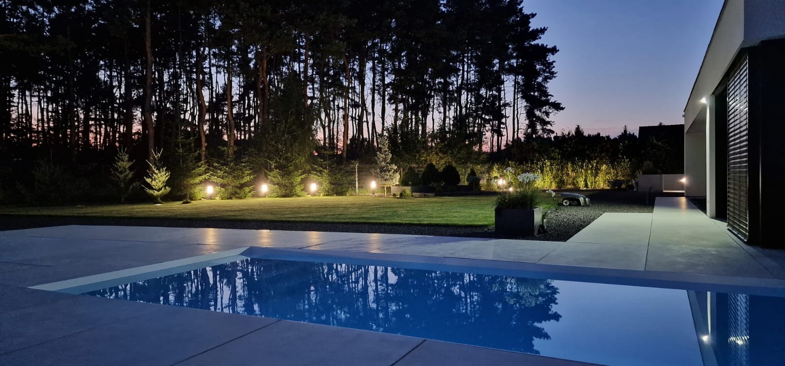Nastrojowe oświetlenie ogrodu z automatyczną kosiarką, widok z perspektywy basenu, w tle las i zachodzące słońce.
