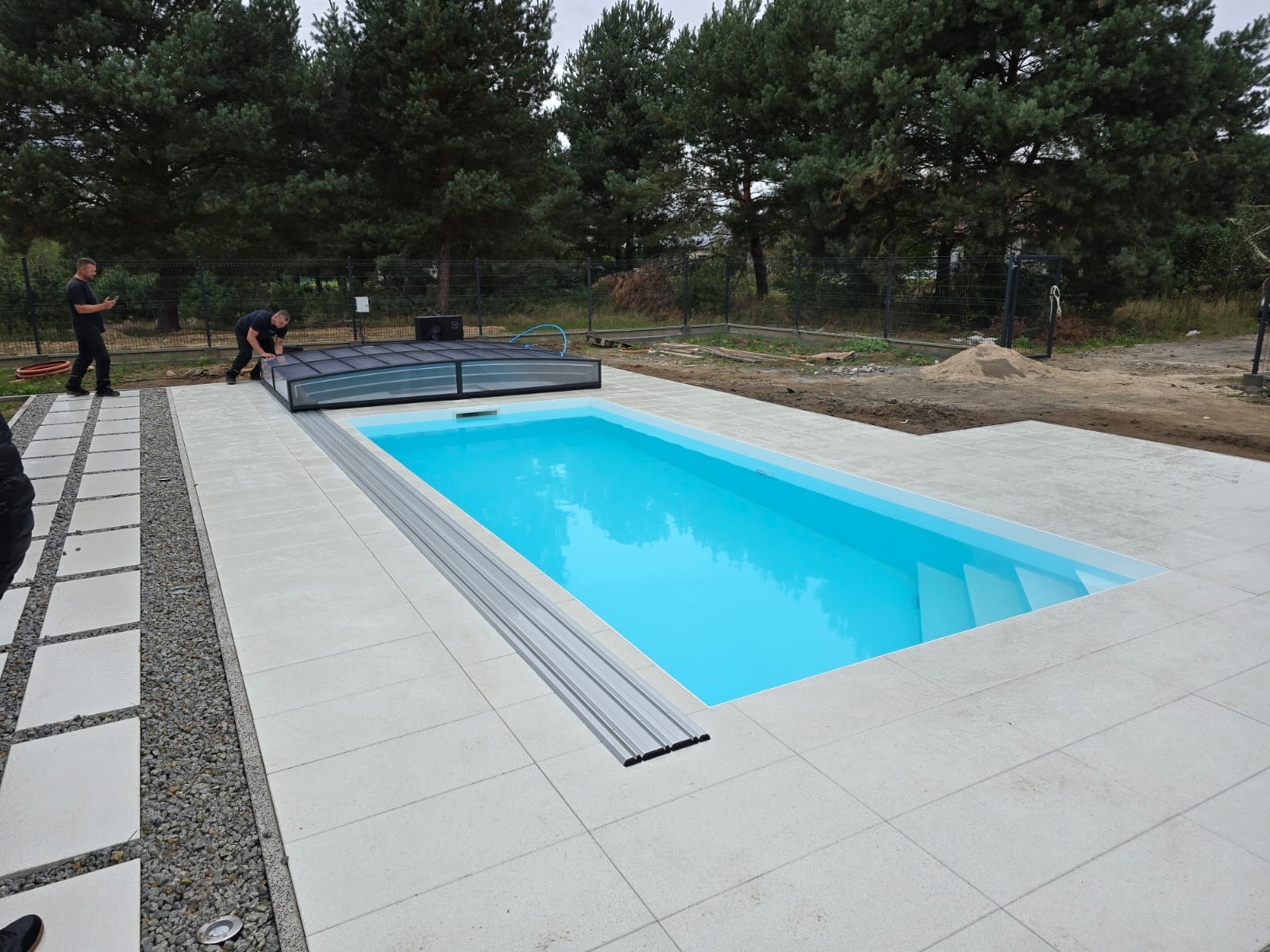 Montaż zadaszenia basenu z turkusową wodą, widoczni dwaj pracownicy w trakcie instalacji, otoczenie wyłożone jasnymi płytami, w tle drzewa.