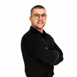 Uśmiechnięty mężczyzna w czarnej koszuli i okularach, z założonymi rękami na białym tle.