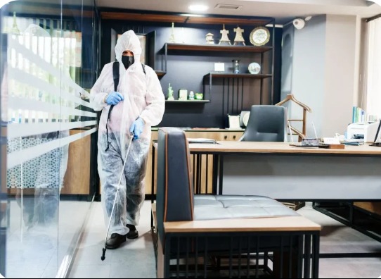 Osoba w kombinezonie ochronnym i masce, rozpyla preparat w biurze z przeszkloną ścianą, widoczny biurko z krzesłem i półki z dekoracjami.
