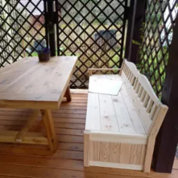 Wnętrze altany z drewnianym stołem i ławką ze schowkiem, widok na kratownicę i roślinność na zewnątrz.