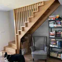 Drewniane schody z ażurową balustradą, zlokalizowane w pomieszczeniu z szarym fotelem i regałem z książkami, w dolnym rogu kadru widoczny pies rasy border collie.