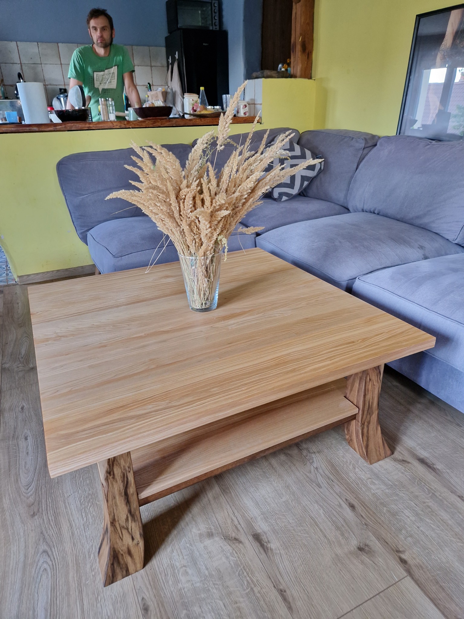 Stolik kawowy z jasnego drewna o kwadratowym blacie i rzeźbionych nogach, na blacie szklany wazon z suszonymi trawami, w tle szara sofa i osoba w kuchni.