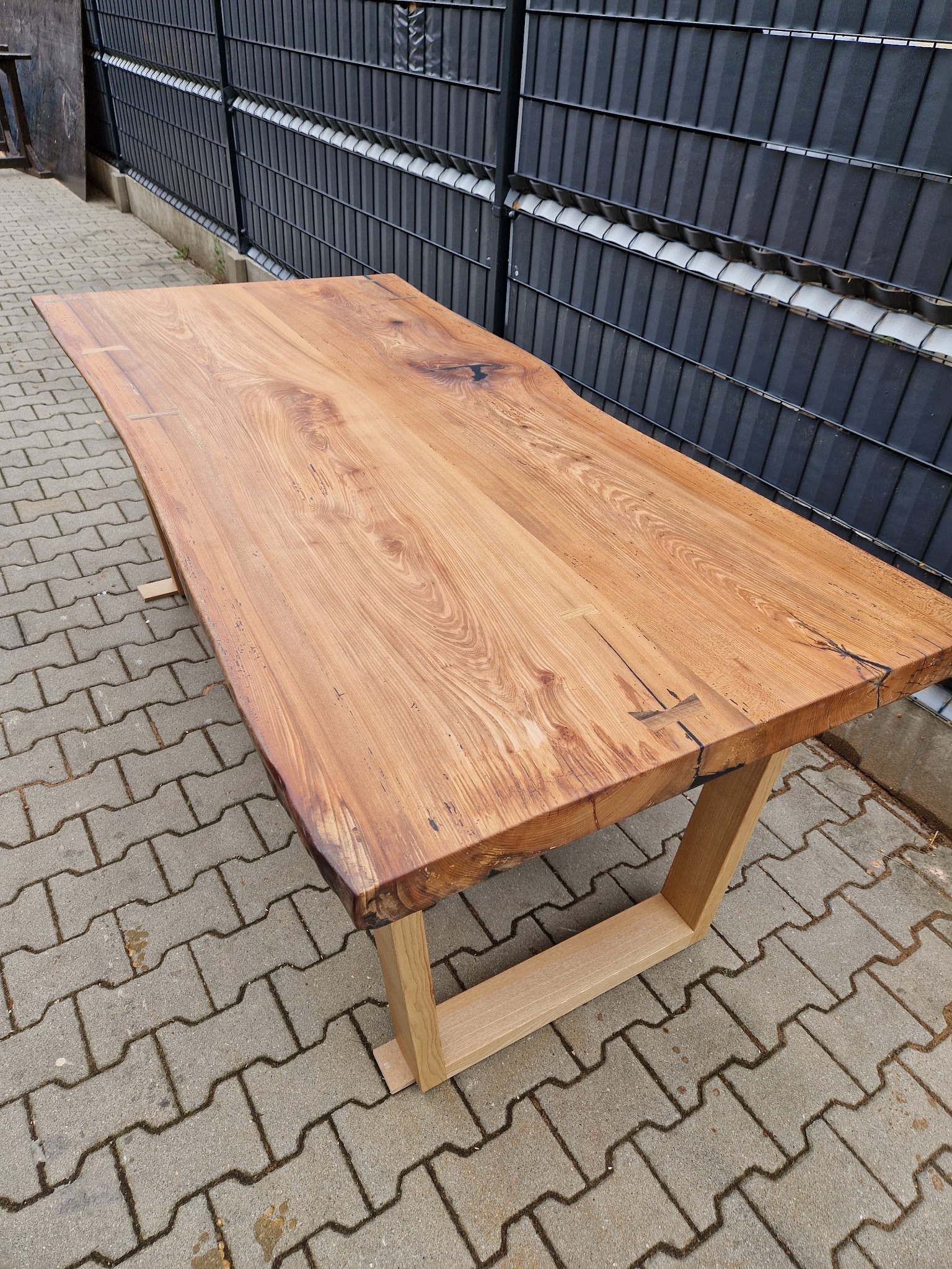 Stół z blatem z litego drewna o nieregularnych krawędziach, osadzony na jasnych, drewnianych nogach o prostokątnym przekroju, na tle szarej kostki brukowej i metalowego ogrodzenia.