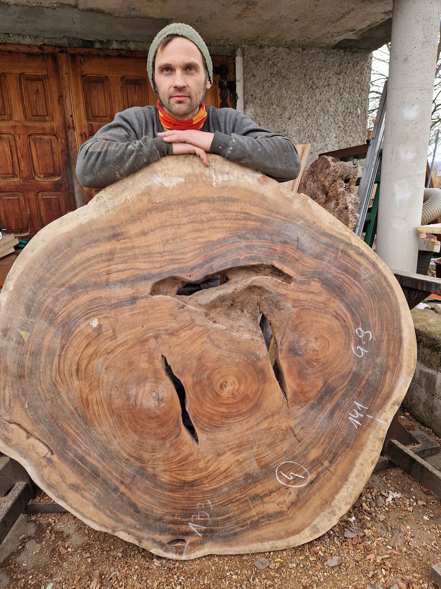Mężczyzna w czapce opiera się o duży, okrągły plaster drewna z widocznymi pęknięciami i oznaczeniami, prawdopodobnie przygotowany do obróbki na blat stołu.