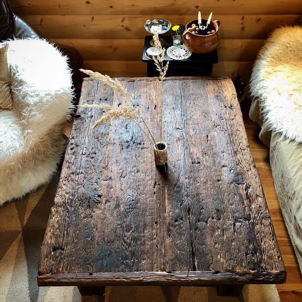 Stary, drewniany stolik kawowy z wyraźnym usłojeniem i śladami użytkowania, w rustykalnym wnętrzu z drewnianą boazerią i elementami dekoracyjnymi, takimi jak suszone trawy w wazonie i futrzane...