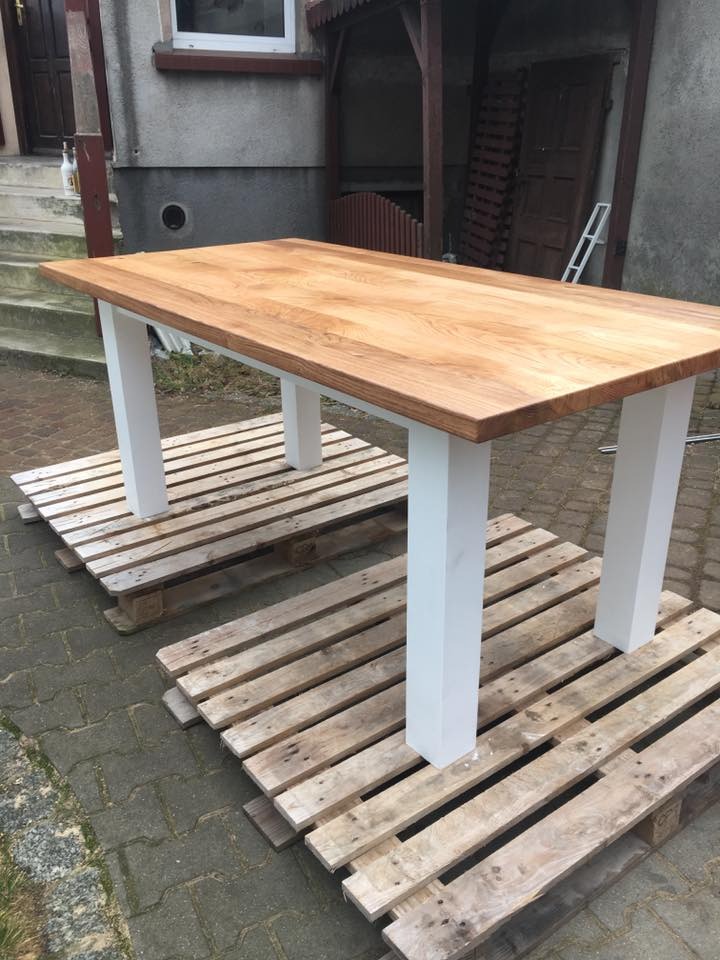 Stół z blatem z jasnego drewna i białymi nogami, ustawiony na dwóch drewnianych paletach przed szarym budynkiem.