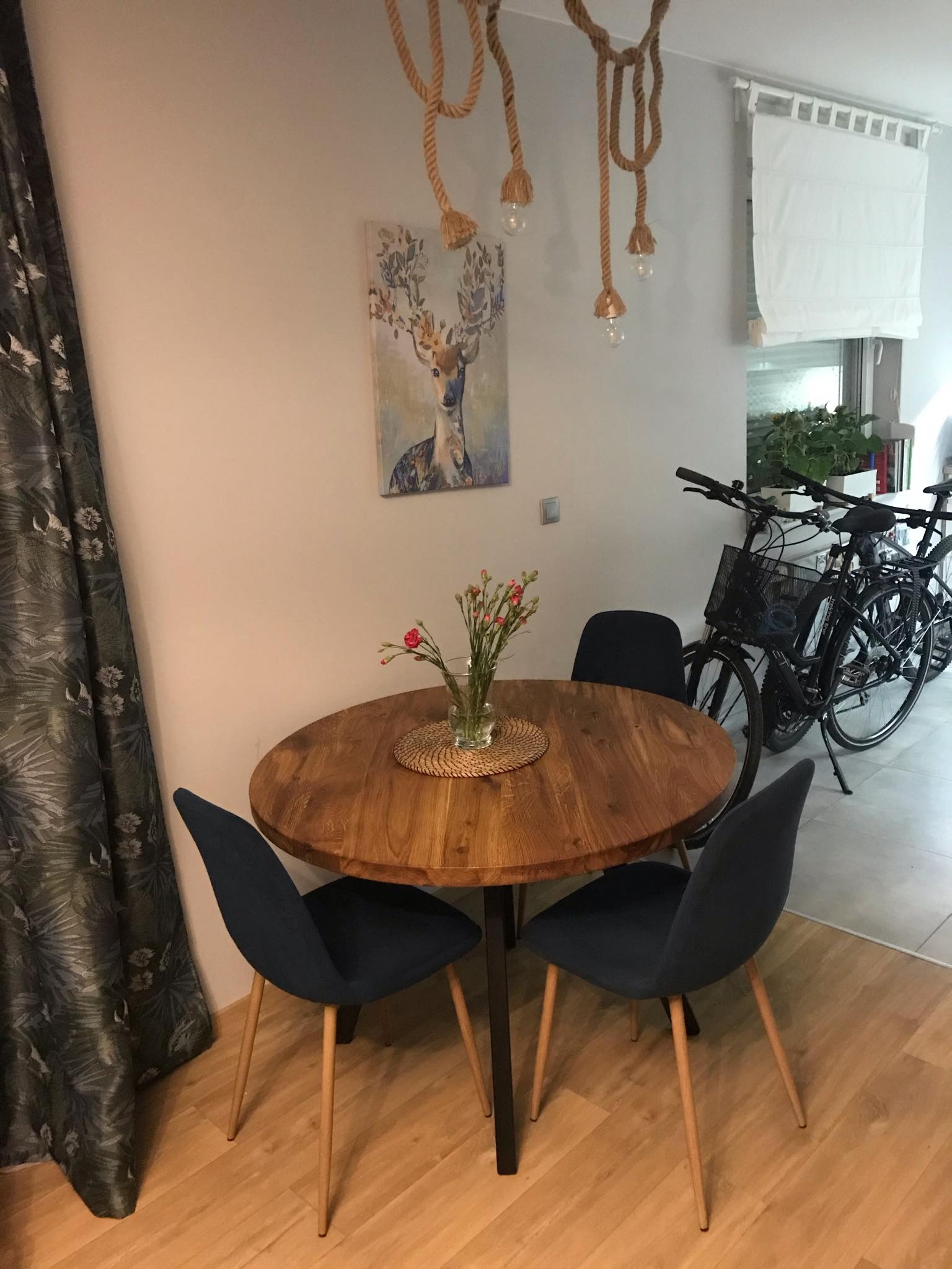 Okrągły drewniany stół na czarnej nodze, otoczony czterema granatowymi krzesłami, z bukietem czerwonych kwiatów w wazonie, wisząca lampa z jutowego sznura i obraz z jeleniem w tle.