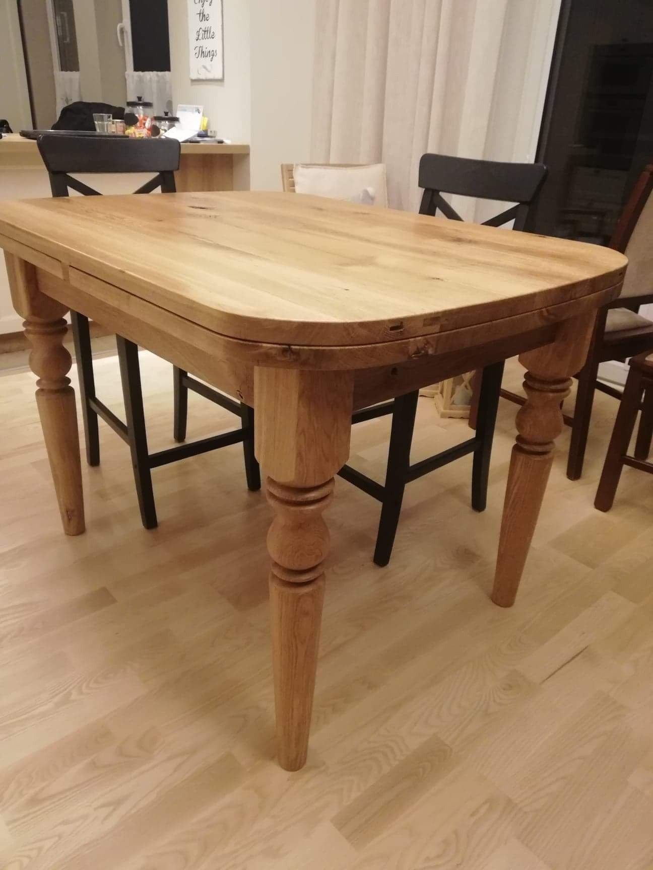 Rozkładany stół z jasnego drewna, z toczonymi nogami, ustawiony na podłodze z paneli; w tle krzesła i zasłony.