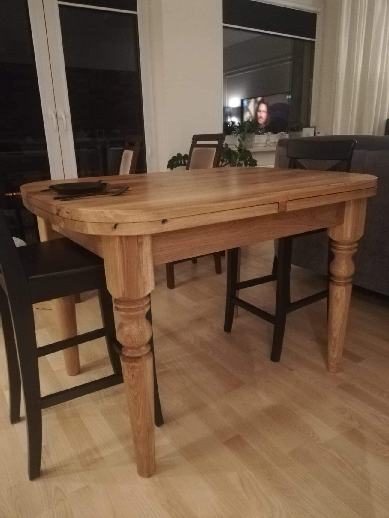 Rozkładany stół z jasnego drewna z toczonymi nogami, otoczony krzesłami w nowoczesnym wnętrzu z parkietem.