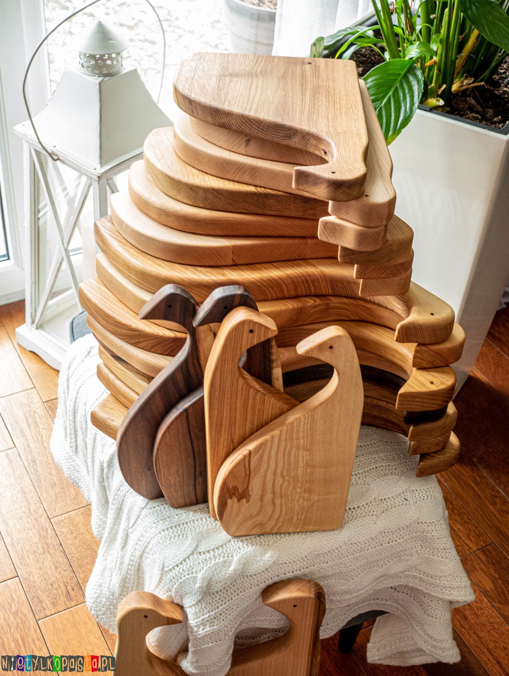 Stos drewnianych desek do krojenia o oryginalnym kształcie z uchwytem, ułożonych na białym, dzierganym pledzie, obok okna i rośliny doniczkowej.